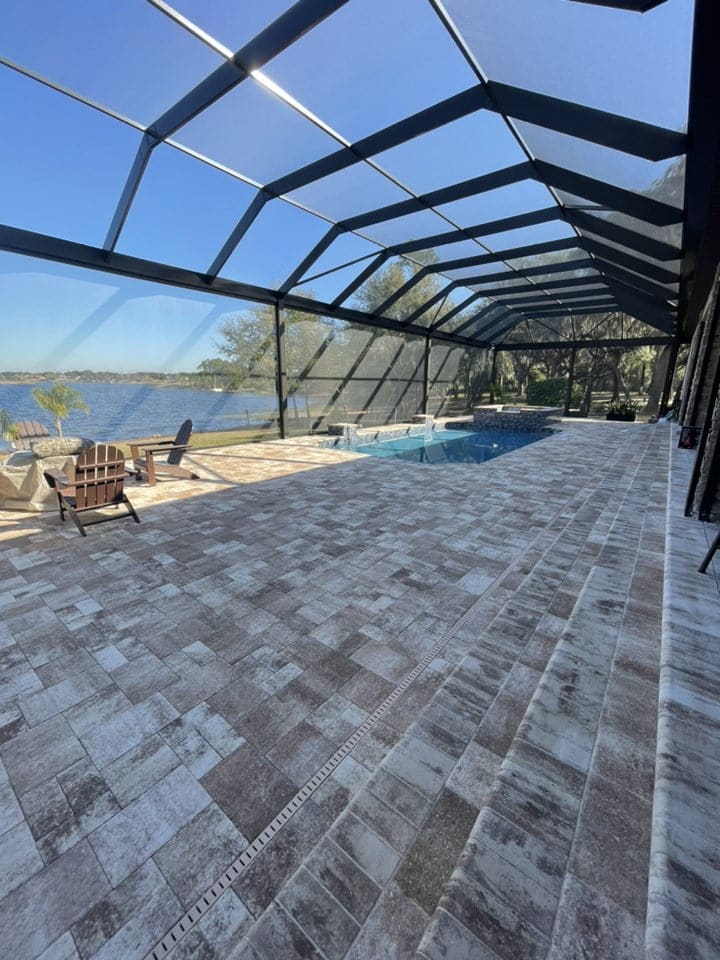 A screened lanai with large paver decking overlooks a lake, with a pool visible in the background.