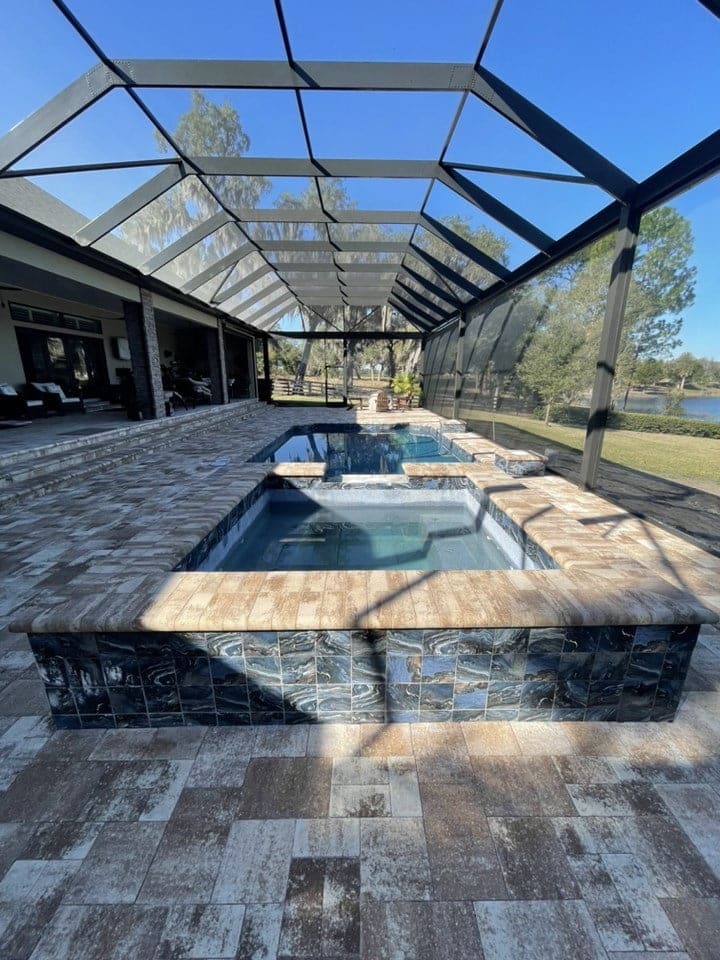 A raised spa with dark mosaic tile sits in front of a pool under a screened lanai overlooking a lake.