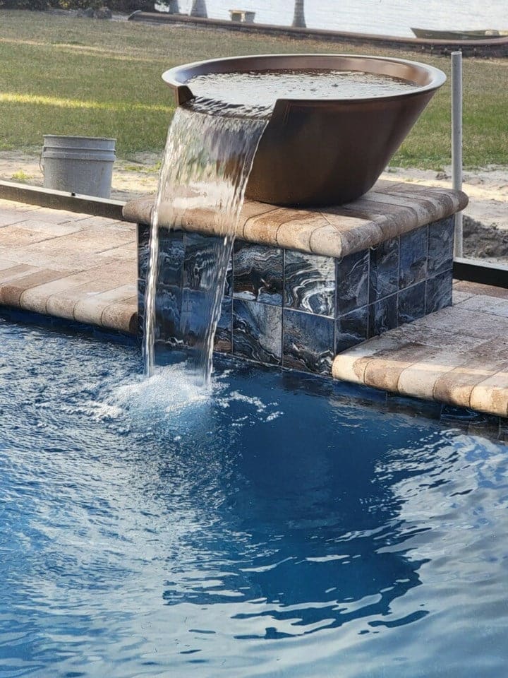 A large copper water bowl spills a sheet of water into a pool over a stone-tiled pedestal near a lakefront yard.
