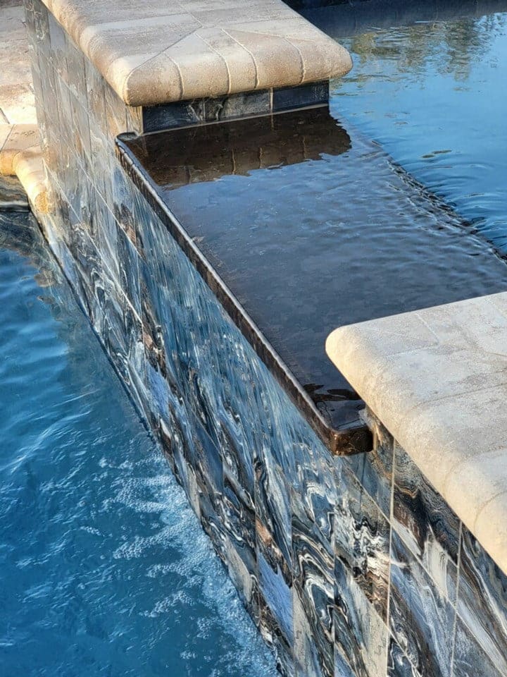 Close-up of a raised spa wall with dark tile spilling water over the edge into the pool below.