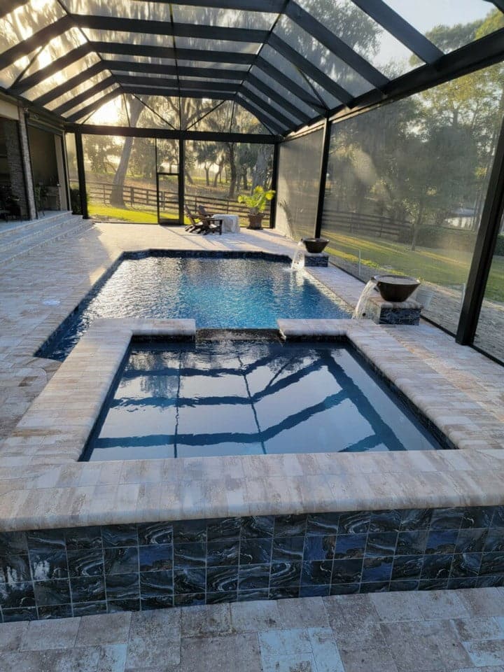 A rectangular pool with an elevated square spa tiled in dark mosaic, under a screened patio enclosure with pastureland visible beyond.