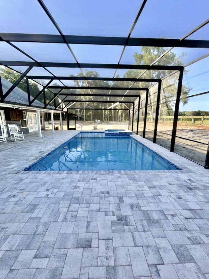 A rectangular vinyl pool with an attached raised spa under a large screened enclosure with paver decking.