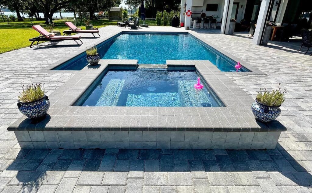 A raised rectangular spa with paver coping sits in front of a rectangular pool overlooking a lake.