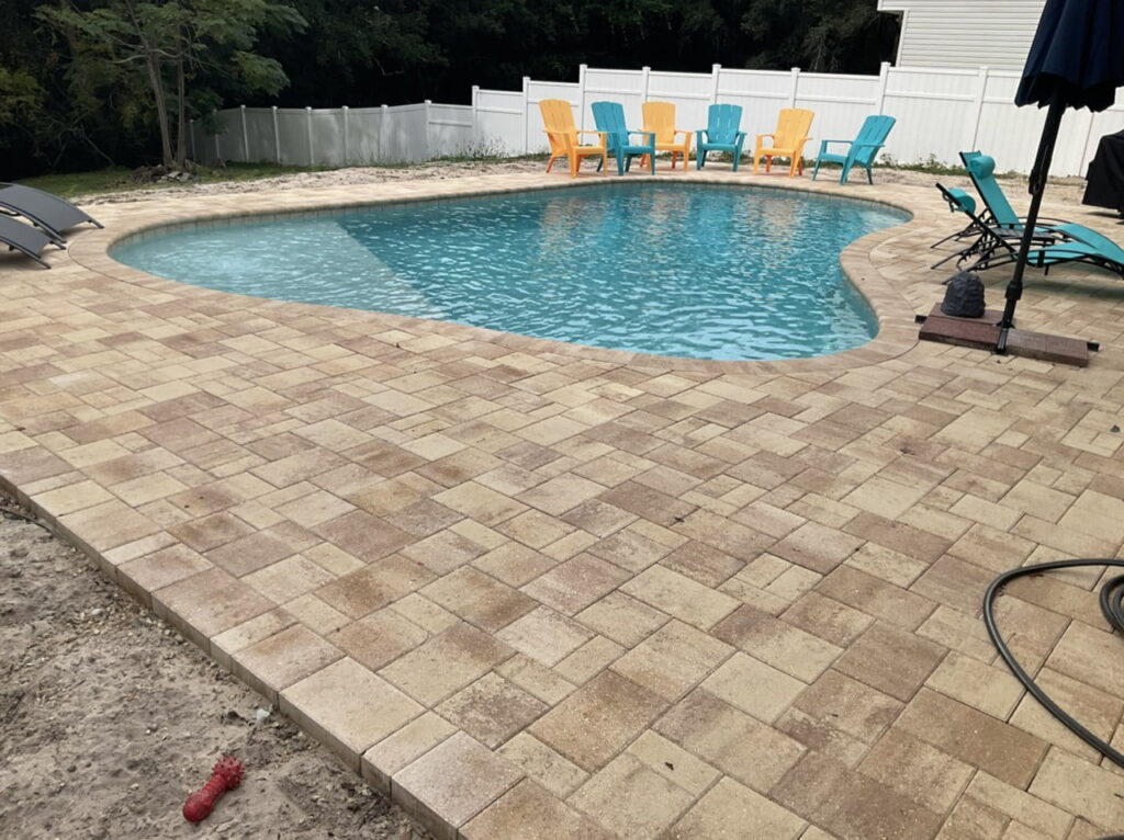 A freeform kidney-shaped pool with turquoise water surrounded by a large paver patio and colorful outdoor chairs.