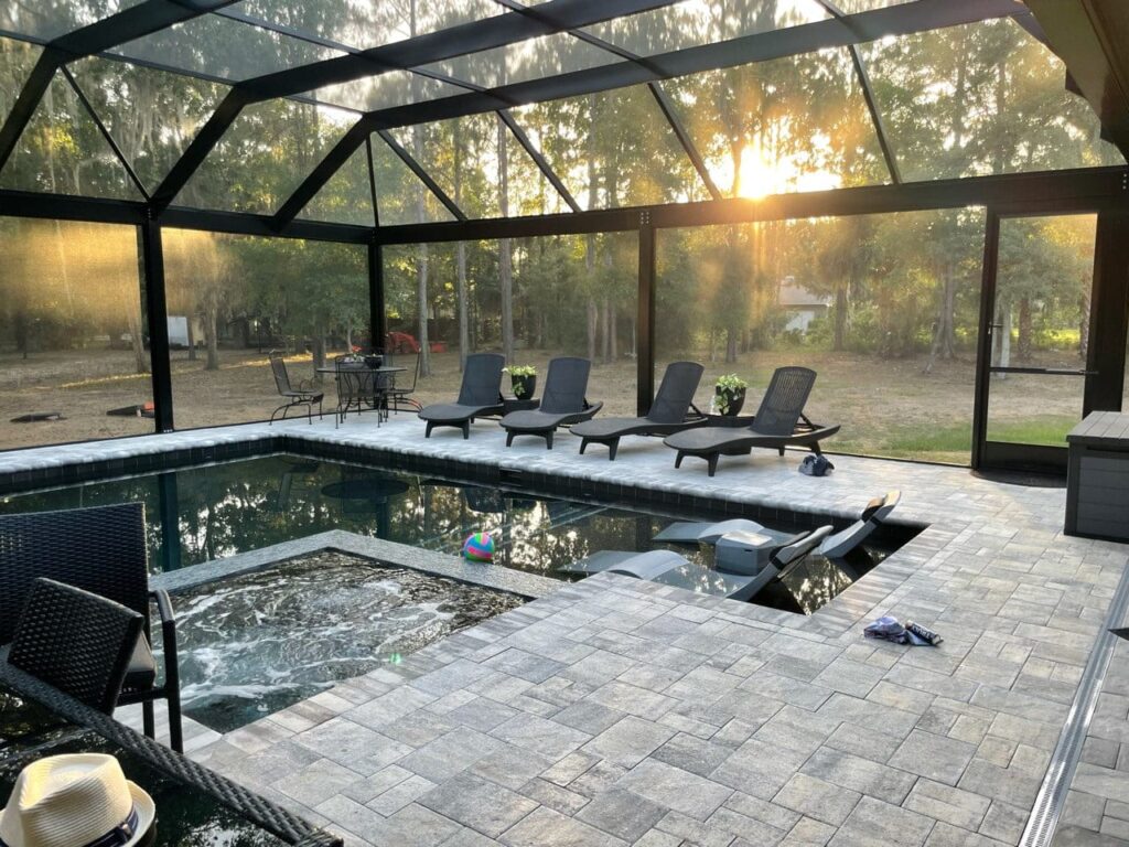 A screened backyard pool with an attached spa and paver deck at sunset, surrounded by trees.