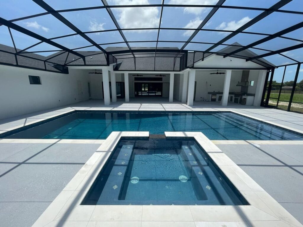 A rectangular pool with a raised spillover spa sits under a large screen enclosure attached to a modern house.