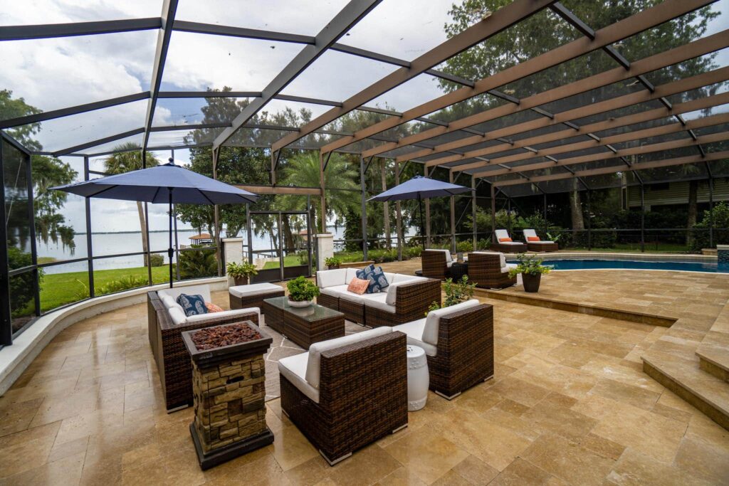 A large screened lanai with wicker lounge furniture and a fire pit, with a pool and lake view in the background.