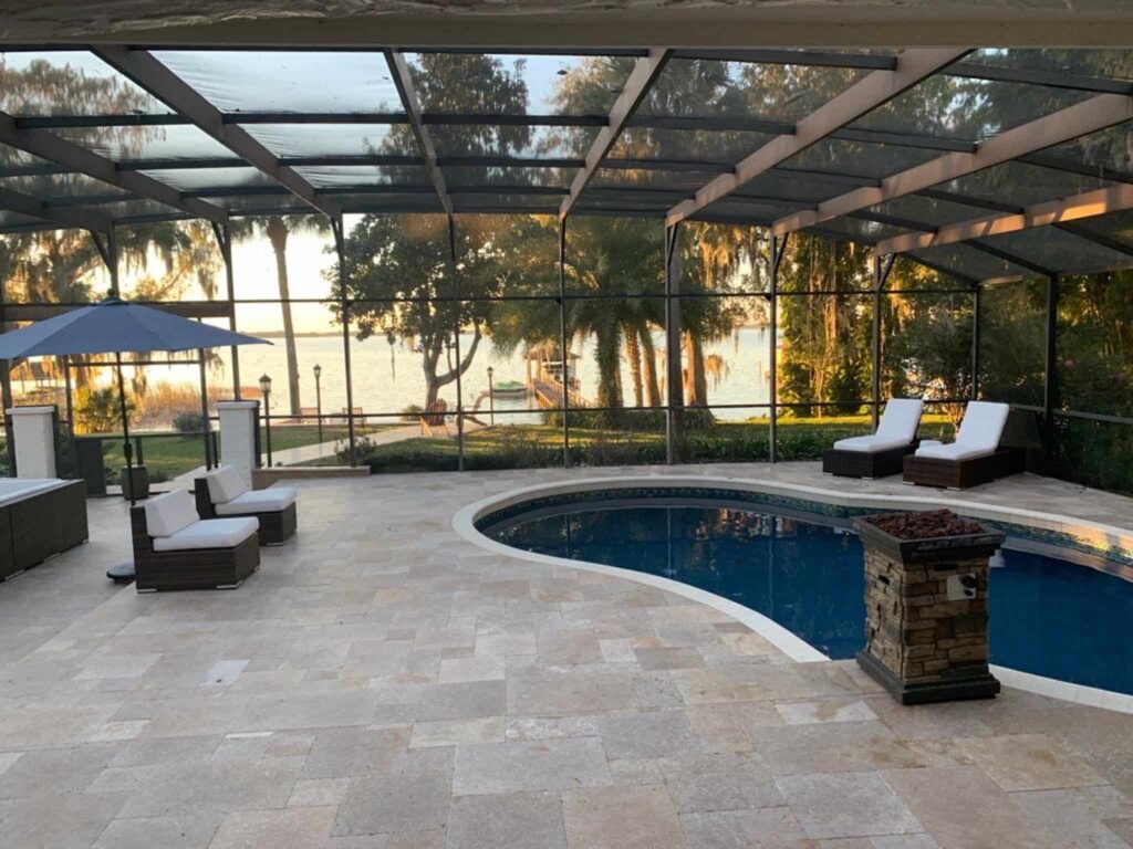 A screened lanai with travertine deck overlooks a kidney-shaped pool and a lake at sunset.