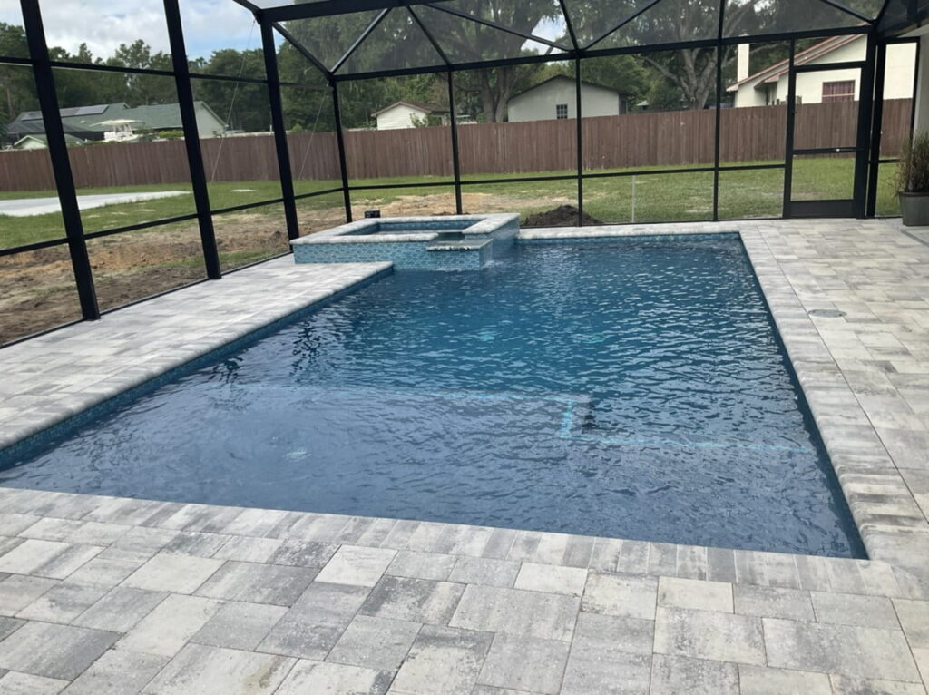A rectangular pool with a raised spa in a screened backyard enclosure with paver decking.