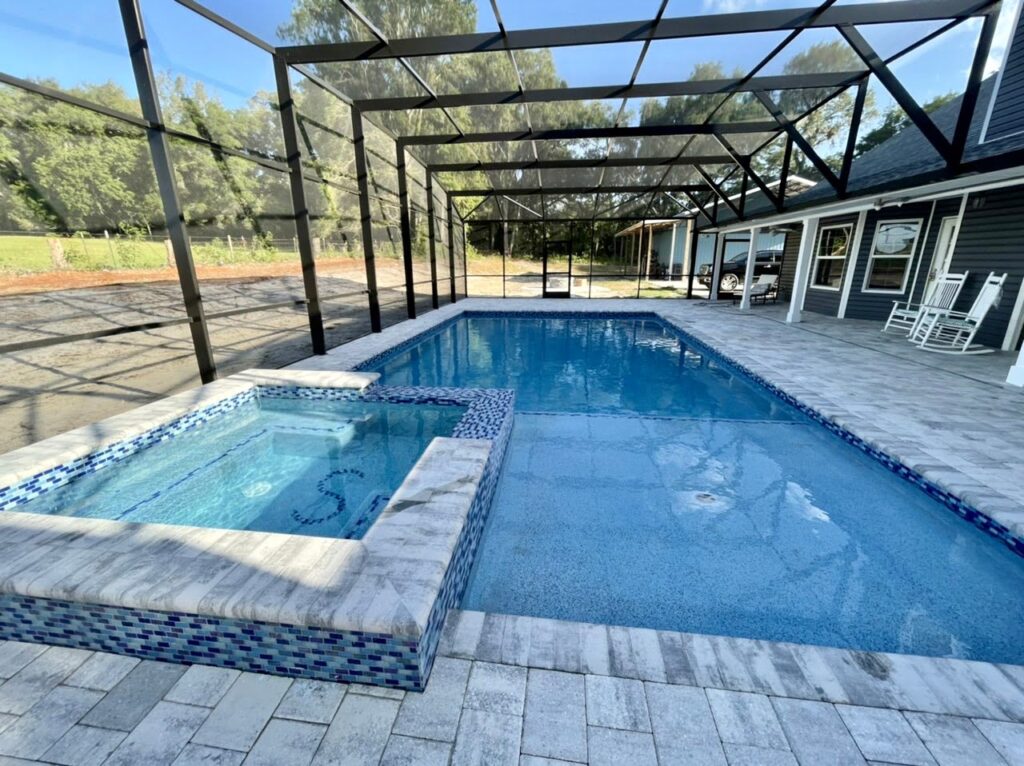 A rectangular gunite pool with a raised tiled spa under a large screen enclosure beside a dark house.
