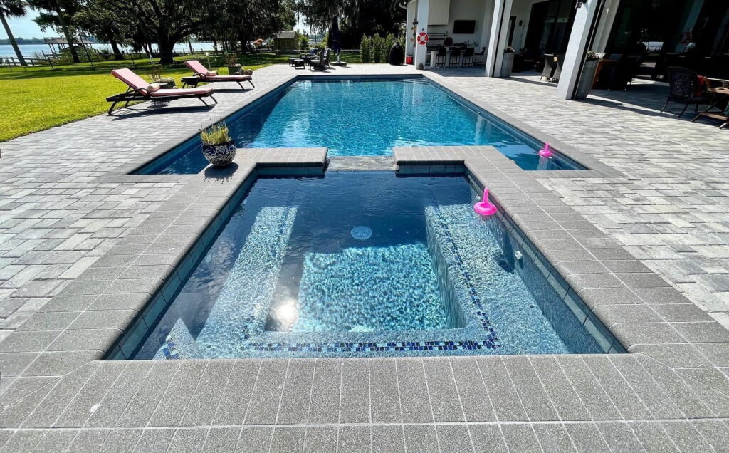 A tiled in-ground spa with steps sits in the foreground of a paver deck, with a rectangular pool and lake view behind.