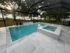 A rectangular pool with a raised tiled spa and sheer descent water feature under a screened enclosure overlooking a lake at dusk.