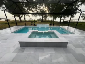 Rectangular pool with a raised square spa on a lakefront lot under a screened enclosure at dusk.