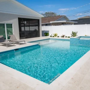 A rectangular backyard pool with a raised spa in the corner, light stone decking, and a white privacy fence under a covered lanai.