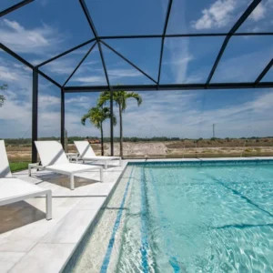 A screened lap pool with blue tile accents and jets, lounge chairs on a travertine deck, and palm trees beyond.