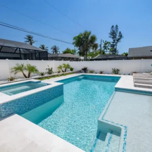 A rectangular pool with a raised tiled spa, sun shelf, and travertine deck in a fenced backyard.