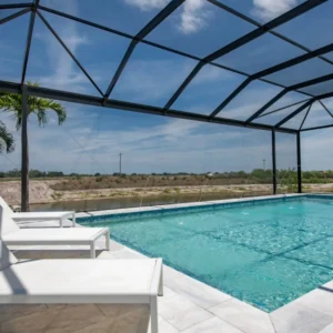 A screened rectangular pool with deck jets overlooks a canal, with white loungers on a travertine patio.