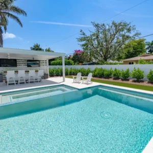 A rectangular backyard pool with a raised spa in front of a covered outdoor kitchen and bar area, surrounded by a white privacy fence and landscaping.