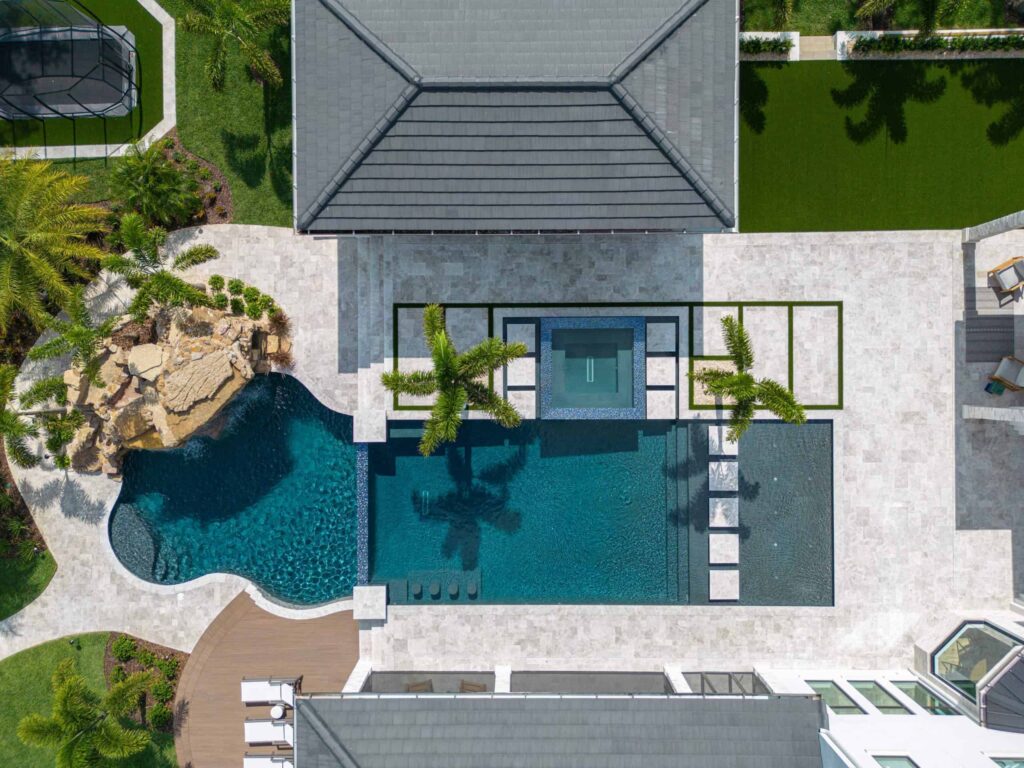 Aerial view of a large freeform pool with a raised spa, natural rock waterfall, and stone paver patio beside a house.