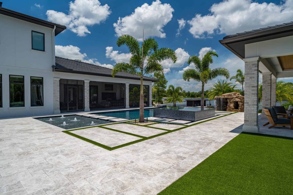 A modern geometric pool with raised spa and fountain jets sits beside a travertine patio overlooking a lake.