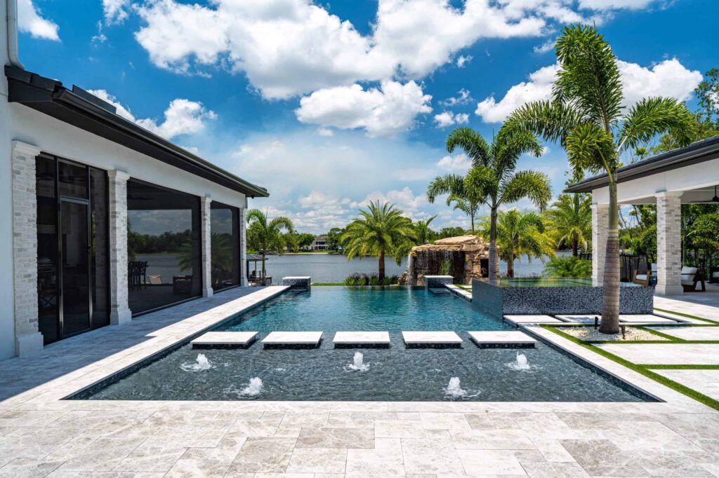 A large infinity-edge pool with floating stepping pads and bubbler jets overlooks a lake, framed by palm trees and travertine decking.