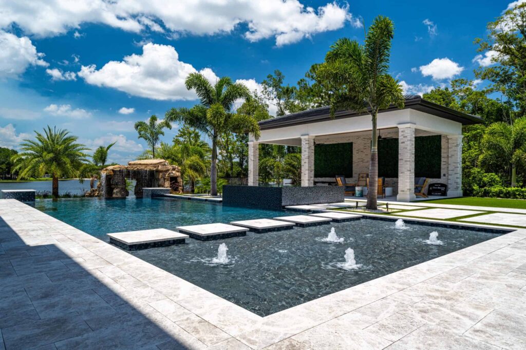 A large multilevel geometric pool with fountain jets, stepping stone pads, and a rock waterfall beside a cabana overlooking a lake.