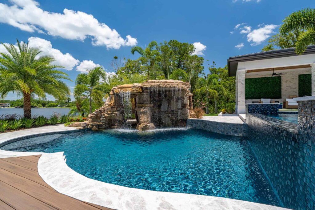 Freeform pool with a large rock waterfall grotto, mosaic tile wall, and lake view behind the home.