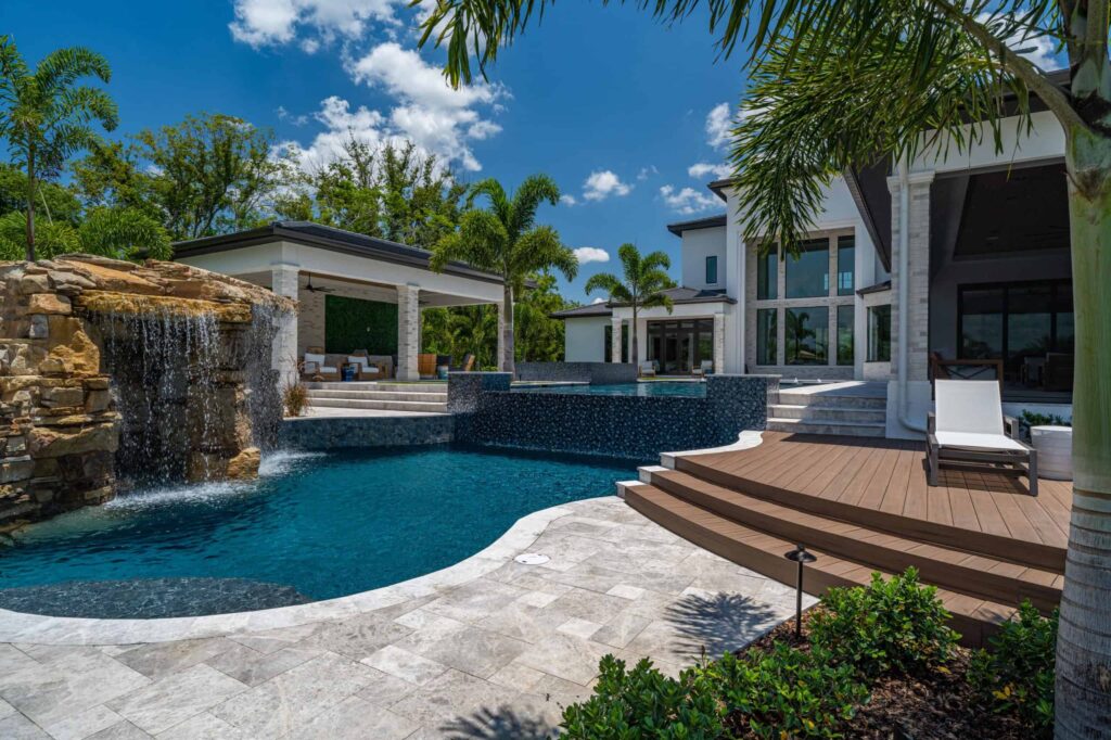 A freeform pool with a rock waterfall sits beside a wood deck behind a large modern home.