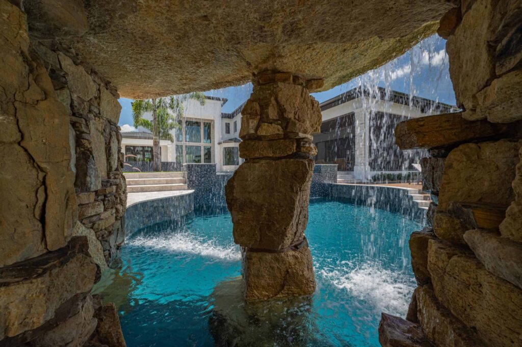 View through a rock grotto with cascading waterfalls into a pool with a large home in the background.