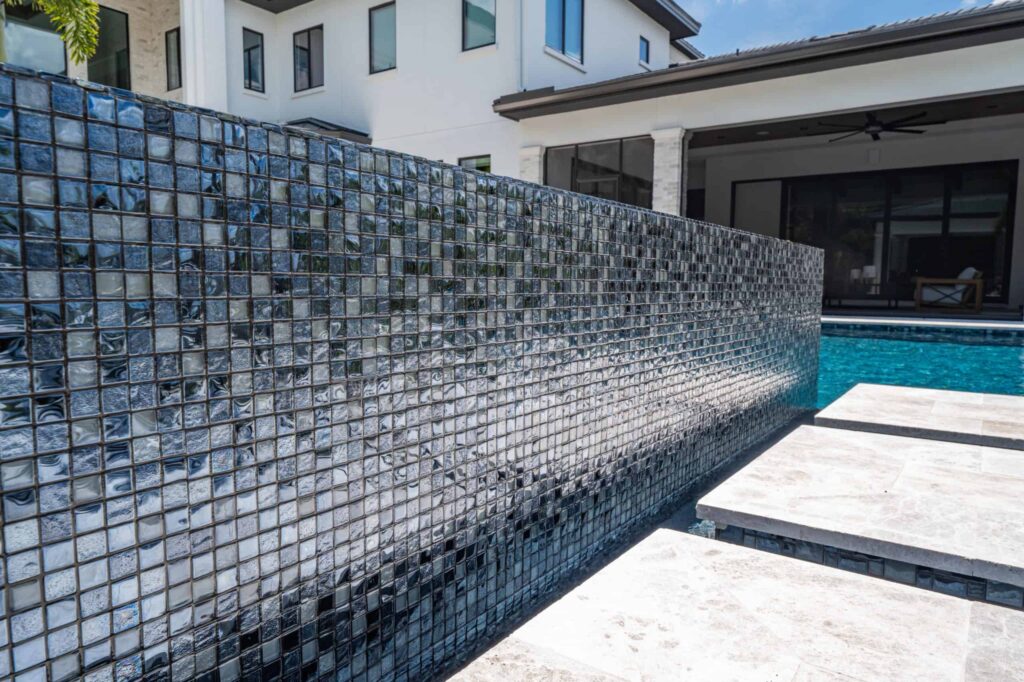 A glass mosaic tile accent wall runs alongside a pool with travertine decking in front of a modern home.