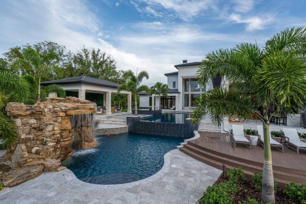 A freeform backyard pool with a raised spa and large rock waterfall in front of a modern white two-story home.