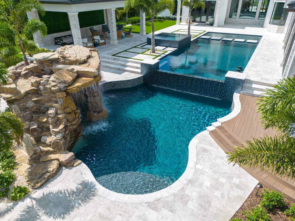 Aerial view of a large freeform pool with raised spa, rock waterfall, and travertine deck beside a modern home.