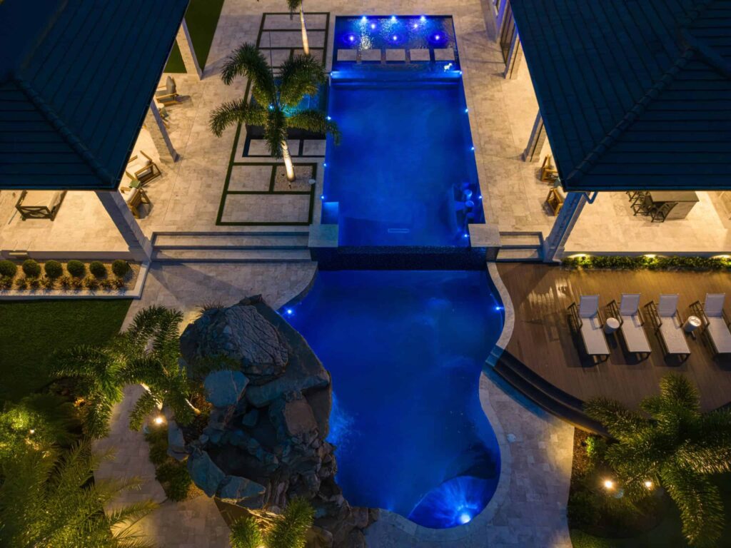 Aerial night view of a freeform pool with a raised spillover spa, boulder waterfall feature, and blue LED lighting between two houses.