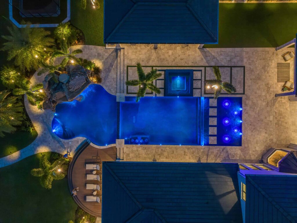 Aerial night view of a freeform pool with raised spa, rock waterfall, and blue LED lighting beside a stone patio.