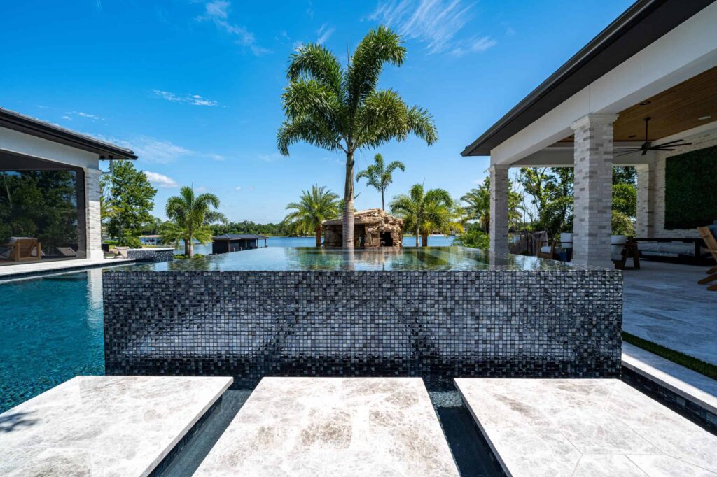 An infinity-edge pool with a dark mosaic tile wall and stone stepping pavers overlooks a palm-lined canal.