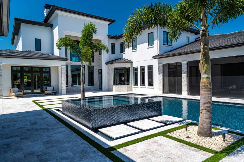 A raised black-tiled spa with floating stepping pads sits beside a rectangular pool in front of a modern white two-story home.