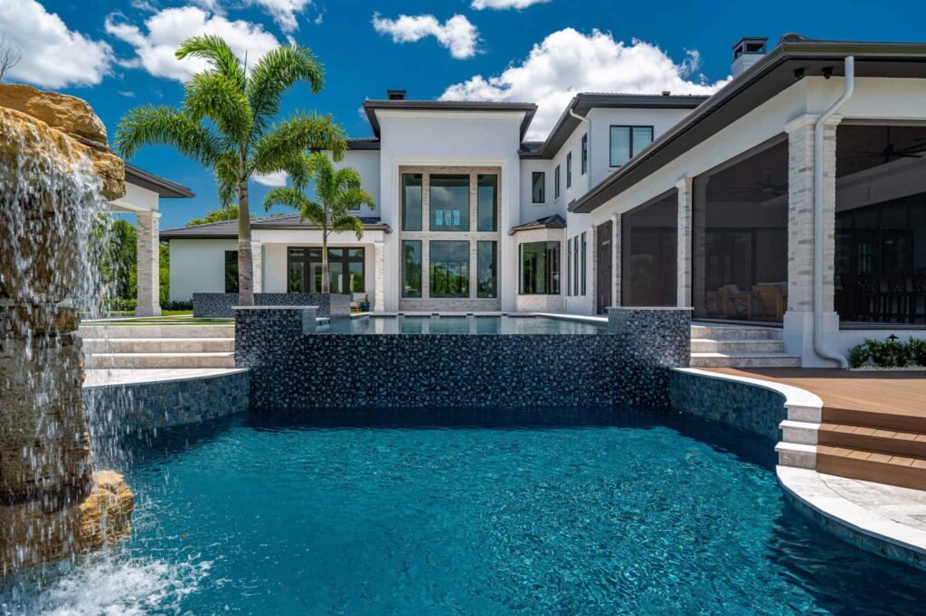 A luxury freeform pool with a raised tiled spa wall and rock waterfall feature behind a large modern white house.