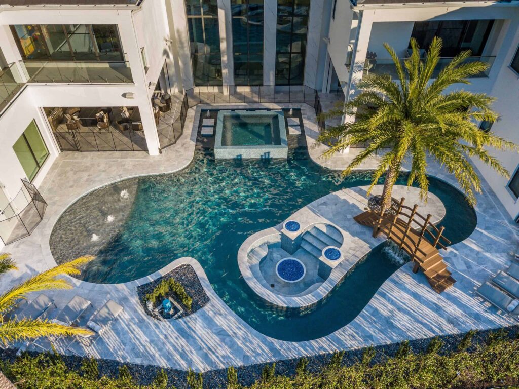 Aerial view of a luxury freeform pool with raised spa, fire bowls, and a wooden bridge to a palm tree island in a courtyard.