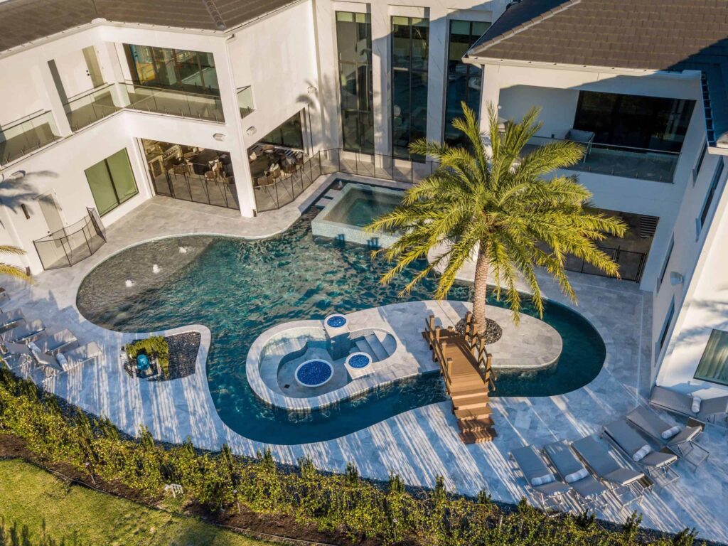 Aerial view of a large freeform pool with a raised spa, wooden footbridge to a palm-tree island, and travertine deck beside a modern white home.