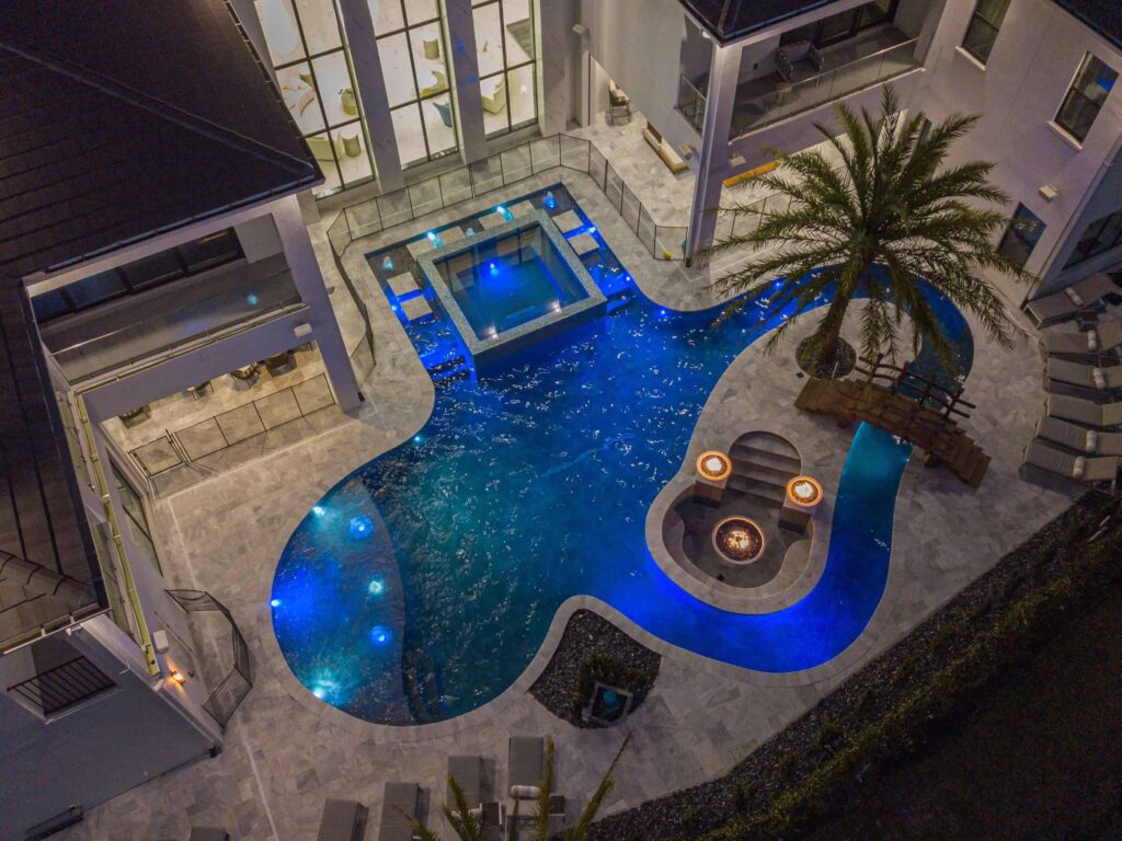 Aerial night view of a freeform pool with a raised glass-tile spa, twin fire bowls, and a lazy river loop around a palm tree.