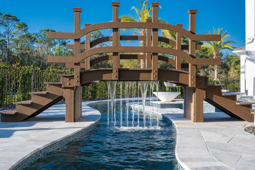 A wooden footbridge arches over a curving pool channel with a rain-curtain water feature falling from its beam.