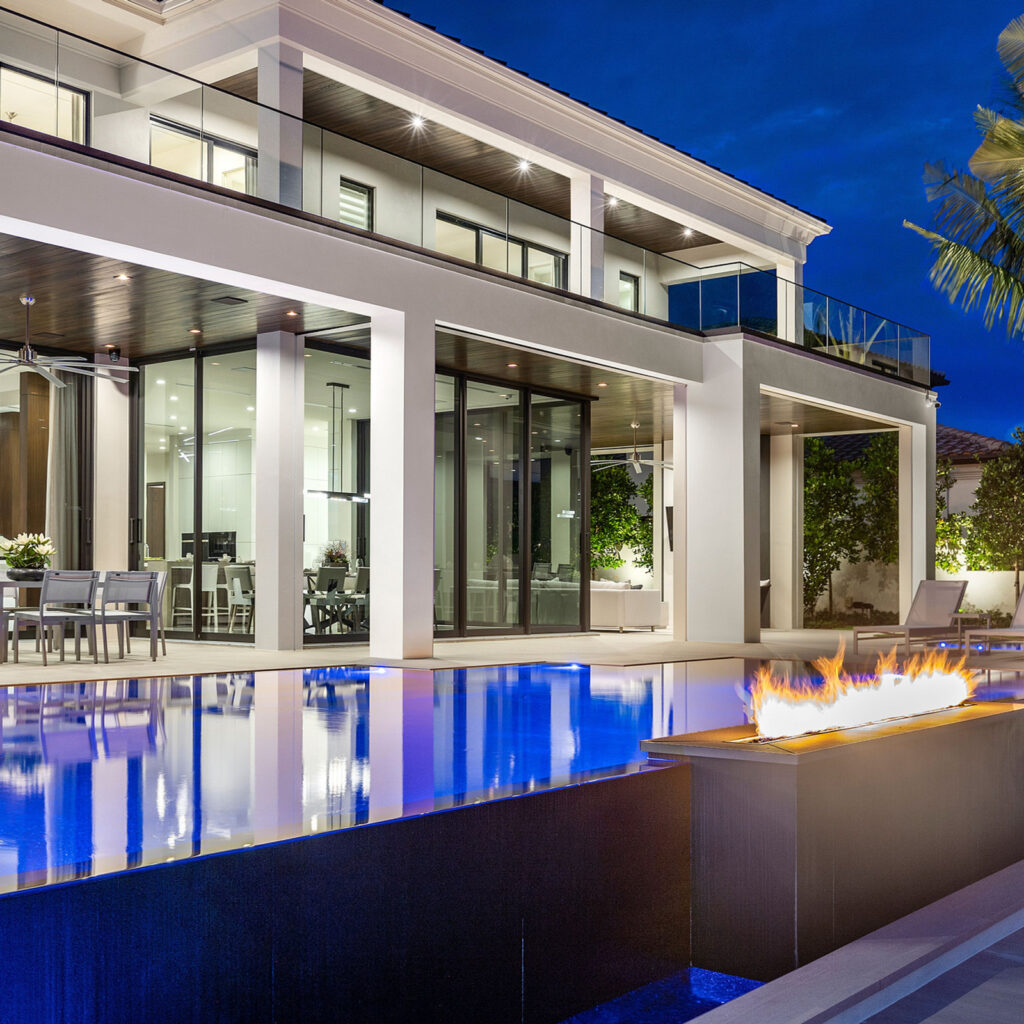 A modern luxury home at dusk with an infinity-edge pool and a lit linear fire trough on the deck.