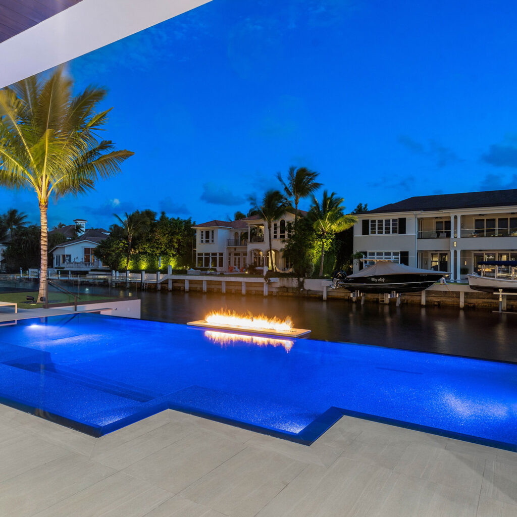 A glowing blue infinity pool at dusk with a floating fire feature and a canal lined with boats and homes beyond.