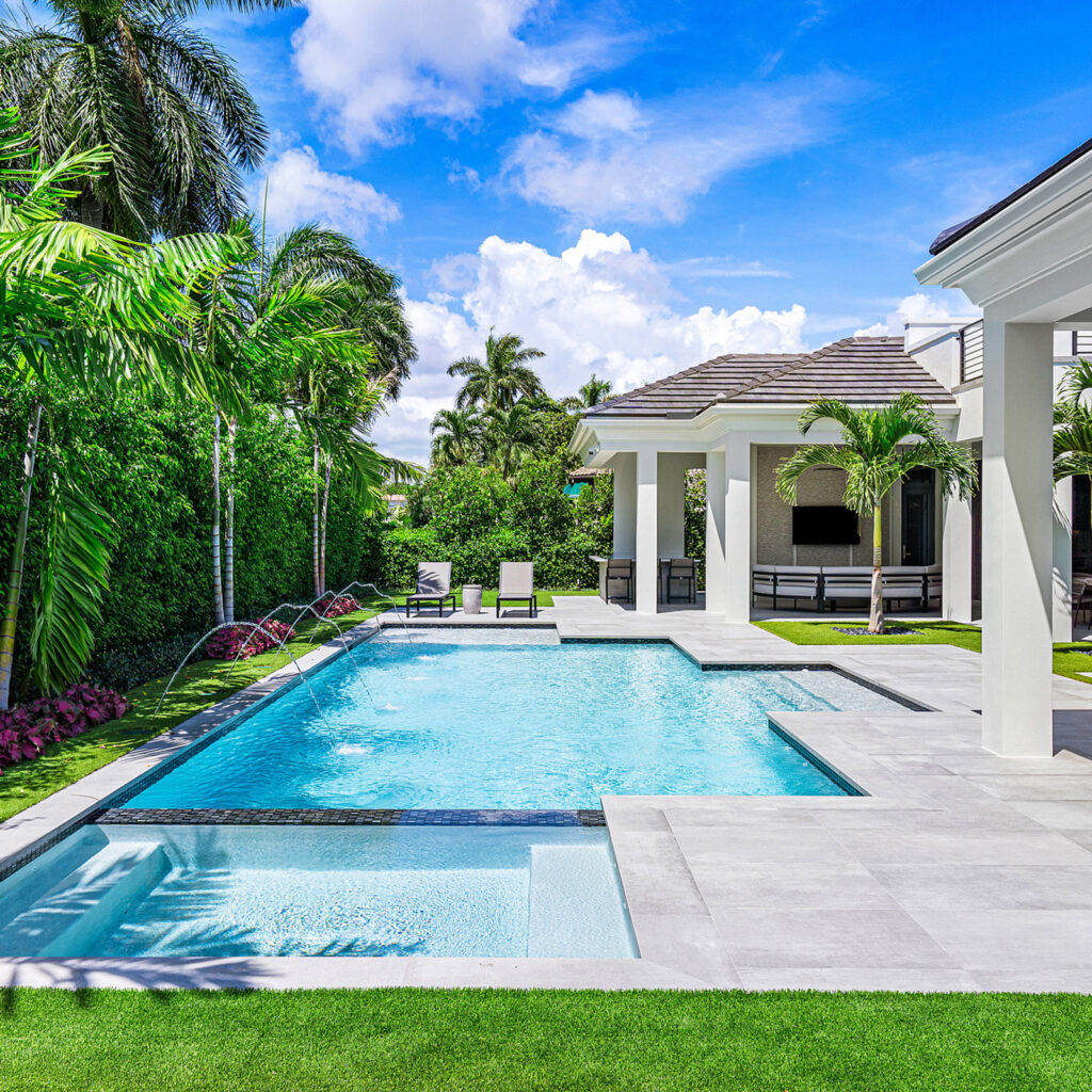 A rectangular backyard pool with a raised sun shelf and deck jets, framed by palm trees and a covered patio.