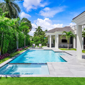 A rectangular backyard pool with a raised sun shelf and deck jets, framed by palm trees and a covered patio.
