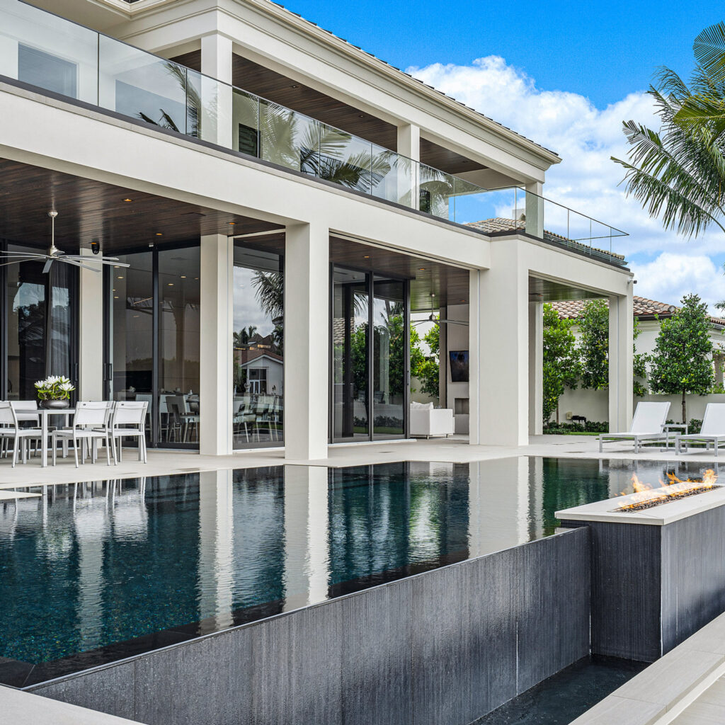 A modern two-story home overlooks an infinity-edge pool with a lit linear fire trough on the patio.
