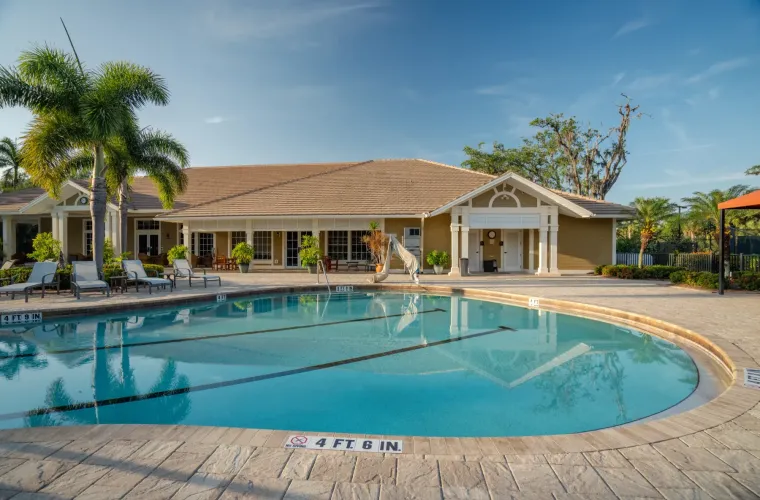 A large community pool with a lane marker in front of a clubhouse with a tiled roof and palm trees.