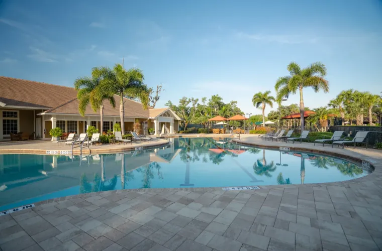 A large freeform community pool surrounded by palm trees, lounge chairs, and a clubhouse building.