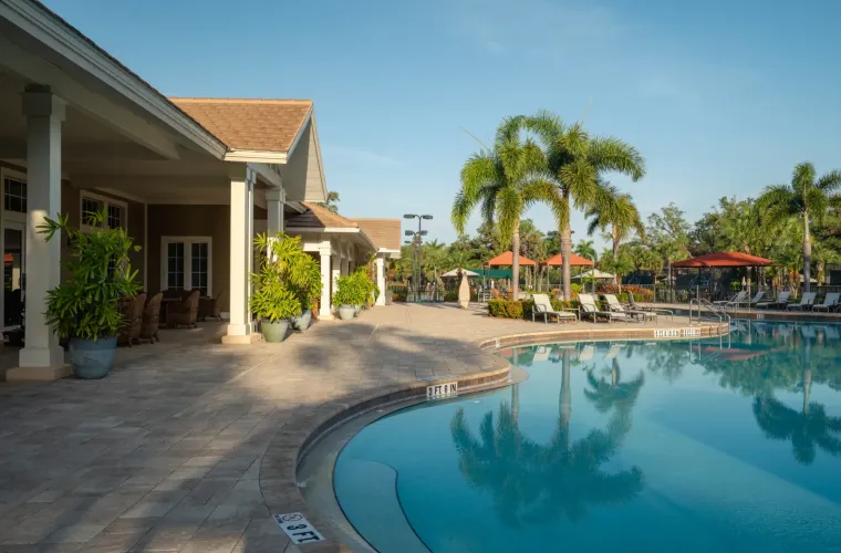 A community clubhouse pool deck with lounge chairs, umbrellas, and palm trees beside a curving pool.