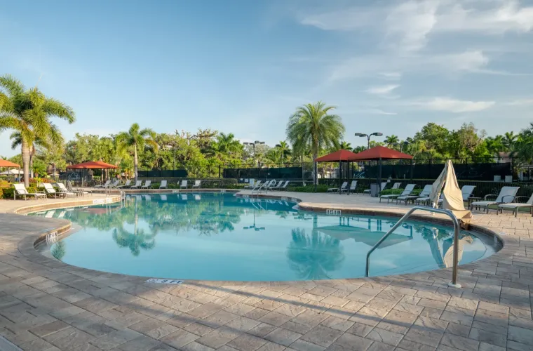 A large freeform community pool surrounded by lounge chairs, red umbrellas, and palm trees under open sky.